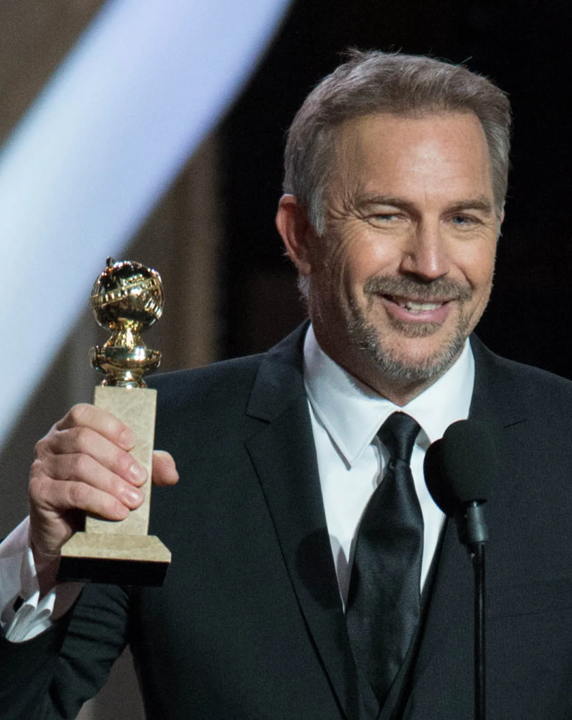 Kevin Costner is wearing a classic black suit and tie while holding a Golden Globe award