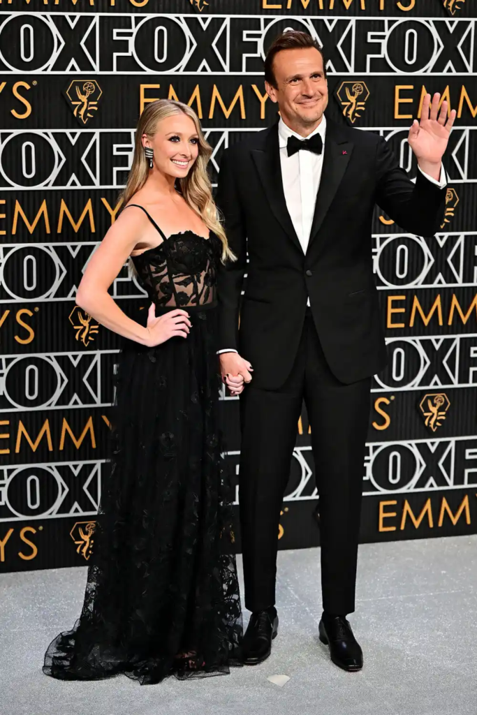 Jason Segel in a classic black tuxedo alongside his fiancée Kayla Radomski, who is wearing an embroidered black gown with a sheer top