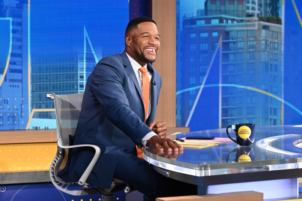 Michael Strahan wears a tailored blue suit paired with a vibrant orange tie while anchoring on the Good Morning America set