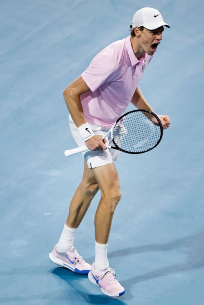 Jannik Sinner wearing a pink Nike polo and white cap while celebrating a point with a racquet in his hand