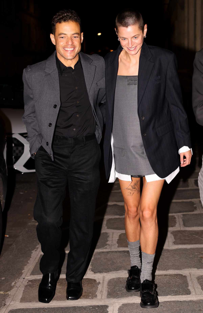 Rami Malek and Emma Corrin leaving a Miu Miu afterparty in Paris, wearing coordinated grey and black tailored outfits.
