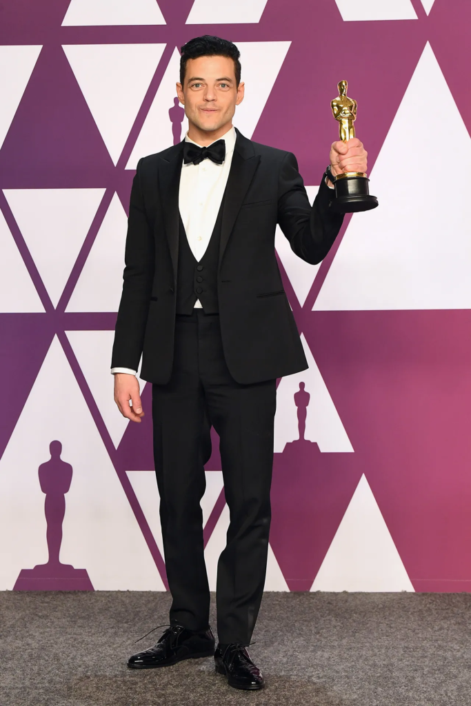 Rami Malek wears a tailored black tuxedo with a crisp white shirt and black bow tie while posing with his Oscar trophy