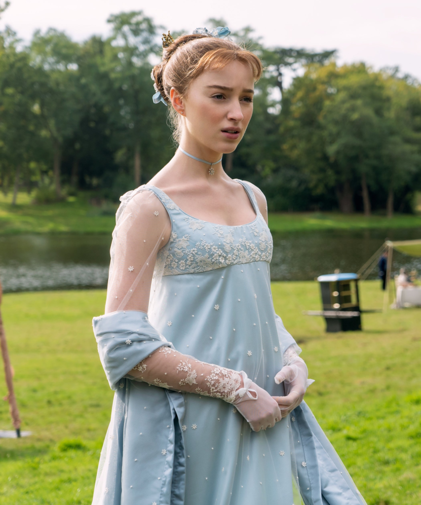 Phoebe Dynevor as Daphne Bridgerton, wearing a signature light blue Regency-style empire gown with sheer long sleeves and delicate embroidery