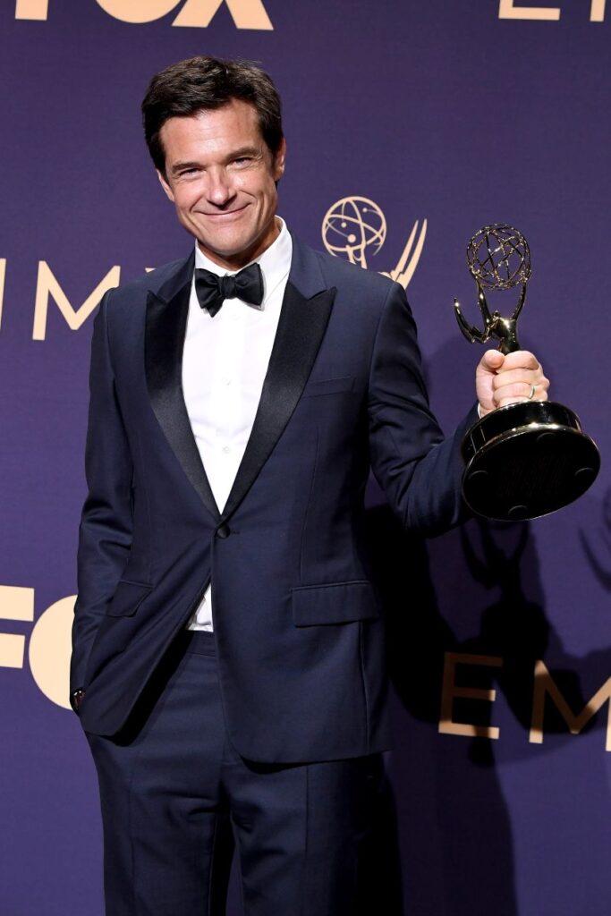 actor Jason Bateman in a dark blue tuxedo posing with an Emmy award