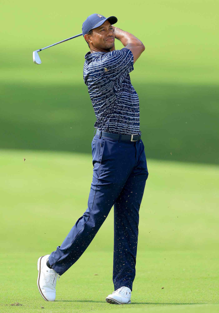 Tiger Woods wearing a blue patterned polo shirt and dark pants while playing a shot on the course