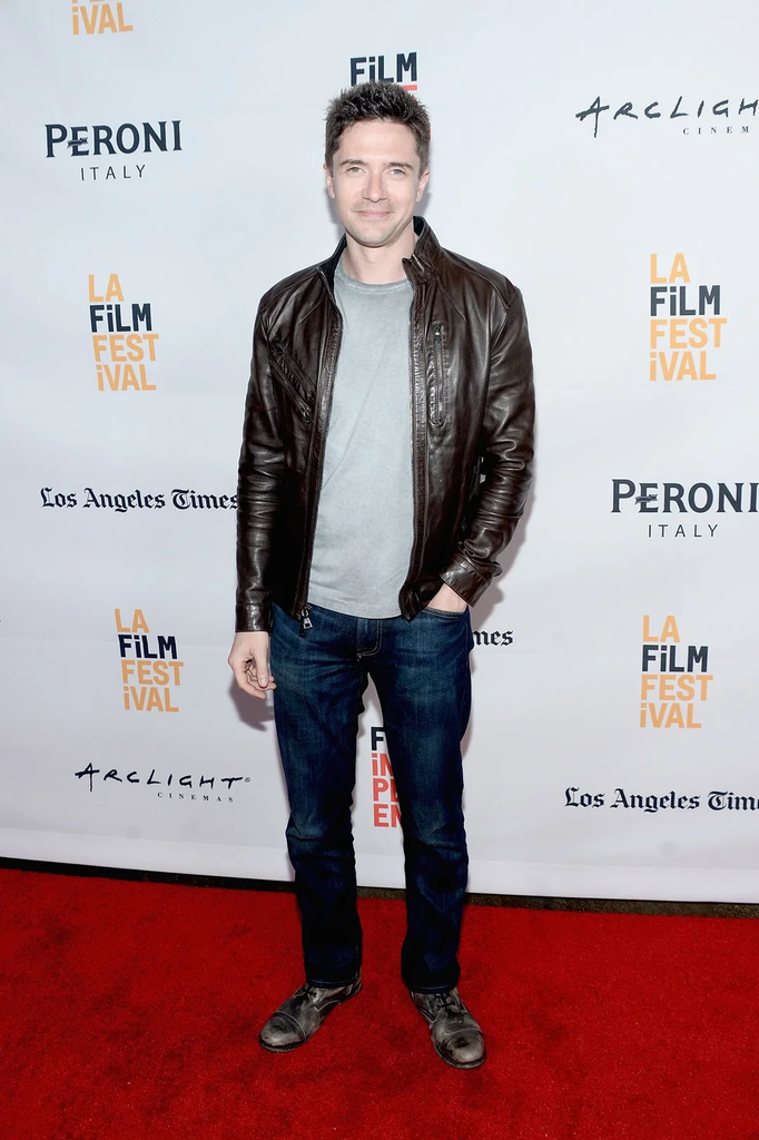 Topher Grace wearing a brown leather jacket and dark jeans on a red carpet