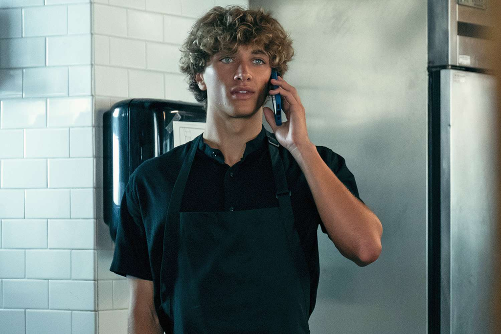 Gavin Casalegno as Jeremiah Fisher in the Prime Video series The Summer I Turned Pretty, appearing in a scene wearing a black work shirt and apron while on the phone