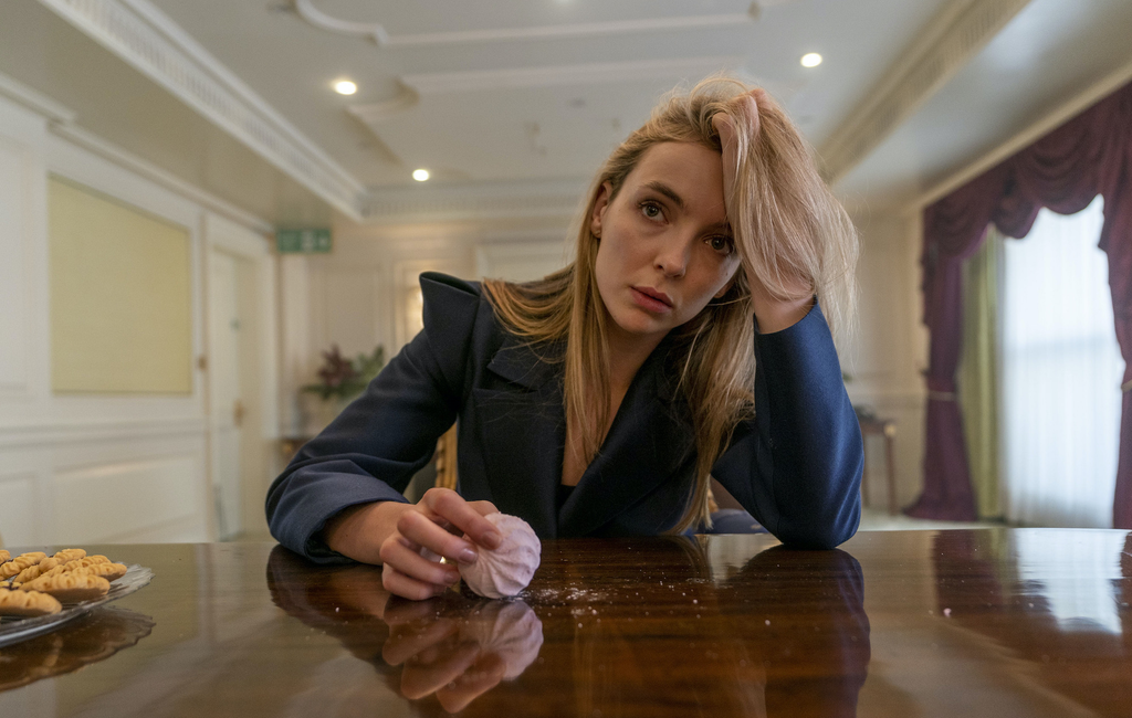 Jodie Comer as the character Villanelle in the television series Killing Eve, resting her head on her hand at a table with a pink marshmallow-like sweet