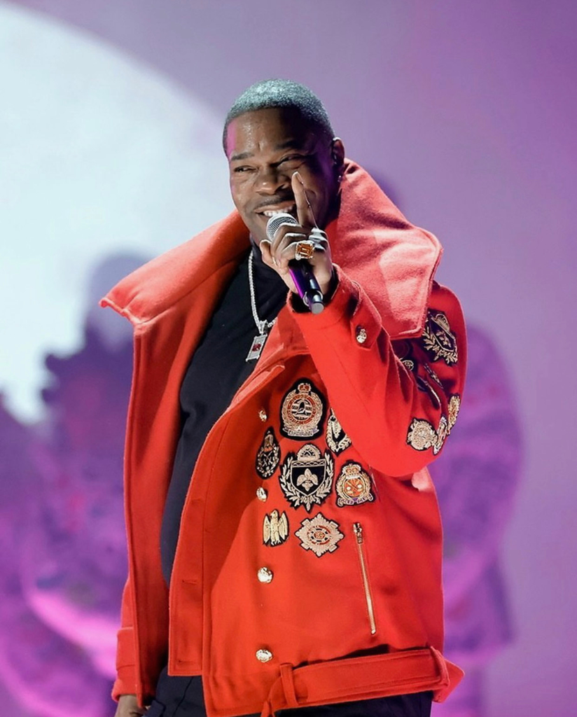 Rapper Busta Rhymes performs on stage wearing a bright red, military-inspired jacket adorned with ornate gold, black, and red heraldic crest and crown patches